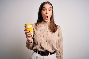 Genç, güzel esmer kadın izole edilmiş beyaz arka planda bir bardak kahve içiyor. Şaşırmış bir yüzle korkmuş ve korku ifadesinden heyecanlanmış.