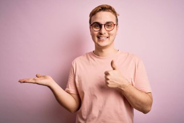 Genç, kızıl saçlı, sıradan bir tişört giyen, pembe arka planda duran, avuçlarını gösteren, baş parmaklarıyla hareket eden, mutlu ve neşeli gülümseyen bir adam.