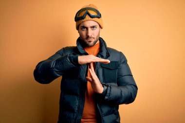 Kar giysileri ve kayak gözlüğü takan sakallı yakışıklı kayakçı elleri ve ciddi yüzüyle jest yapıyor.