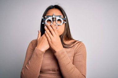 Genç Asyalı optik kız beyaz arka planda optometri gözlüğü kullanarak görme yetisini kontrol ediyor. Ağzı yanlışlıkla ellerle kapatıyor. Gizli konsept.