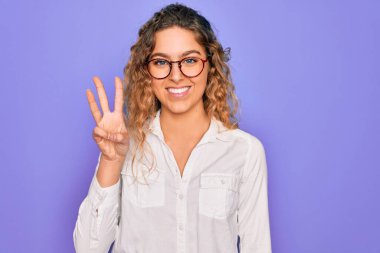 Mavi gözlü genç bir kadın mor arka planda günlük gömlek ve gözlük takıyor. Üç numaralı parmaklarıyla işaret ediyor ve gülümsüyor. Kendinden emin ve mutlu..