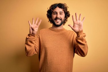 Sakallı, sıradan bir süveter giyen, sarı arka planda duran ve dokuz numaralı parmaklarıyla işaret eden genç ve yakışıklı adam kendinden emin ve mutlu bir şekilde gülümsüyor..