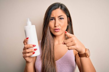 Genç, güzel esmer kadın, izole edilmiş beyaz arka planda elinde bir şişe kremle kamerayı işaret ediyor. Ve sen, el işareti, ön taraftan pozitif ve kendinden emin bir hareket.