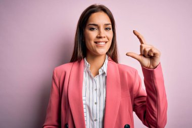 Genç, güzel esmer kadın, pembe arka plan üzerinde duran, izole edilmiş bir ceket giyiyor. Gülümsüyor ve el hareketleriyle küçük beden işareti yapıyor, parmakları görünüyor ve kameraya bakıyor. Ölçü kavramı.