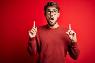 Sakallı, gözlüklü ve süveterli genç adam kırmızı arka planda dikilmiş şaşkın şaşkın yukarı bakıyor, parmaklarıyla işaret ediyor ve kollarını kaldırıyordu..