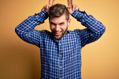 Sarışın, sakallı ve mavi gözlü genç bir işadamı sarı arka planda gömlek giyiyor komik ve çılgın pozlar veriyor parmakları kafasında tavşan kulağı, neşeli bir gülümseme