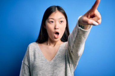 Genç ve güzel Asyalı bir kadın mavi izole edilmiş arka planda dikilen sıradan bir süveter giyiyor. Parmakları önde, ağzı açık, şaşırmış bir ifadeyle işaret ediyor.