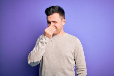 Mor izole arka plan üzerine günlük süveter giyen genç beyaz bir adam pis ve iğrenç bir koku alıyor, katlanılmaz bir koku, parmakları burnunda nefesini tutuyor. Kötü kokuyor.