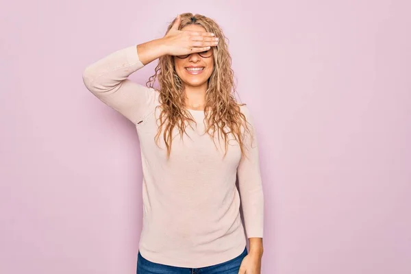 ピンクの背景にカジュアルなセーターと眼鏡を身に着けている若い美しいブロンドの女性は笑顔で 驚きのために目をカバー顔に手で笑っています ブラインドコンセプト — ストック写真