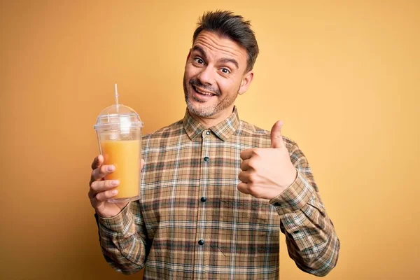 年轻英俊的男人喝着健康的橙汁 用稻草盖住黄色的背景快乐的笑着做着好的手势 用手指竖起大拇指 好的标志 — 图库照片