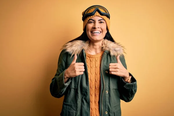 冬の天気の成功のためにスキーグラスとコートを着て若い美しいヒスパニック系の女性は 手で積極的なジェスチャーを行うサイン 親指を笑顔と幸せ 陽気な表情と勝者のジェスチャー — ストック写真