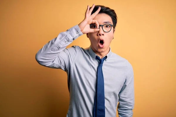 若いハンサムな中国のビジネスマンの眼鏡を着用し 黄色の背景の上にネクタイをOkジェスチャーを行う驚きの顔でショックを受け 目の指を見て 不信仰の表現 — ストック写真