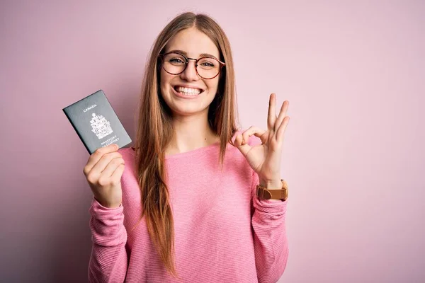 ピンクの背景の上にカナダのカナダのパスポートを保持する美しい赤毛の観光客の女性は指でOkサインをして 優れたシンボル — ストック写真