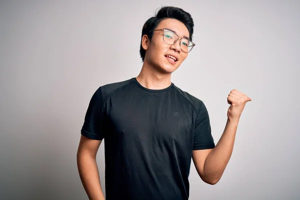Young handsome chinese man wearing black t-shirt and glasses over white background smiling with happy face looking and pointing to the side with thumb up.