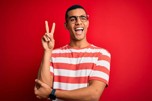 年轻英俊的非洲裔美国男子 身穿休闲条纹T恤 戴着眼镜 高兴地微笑着 用手指在镜头前眨眼示意胜利 第二点 — 图库照片