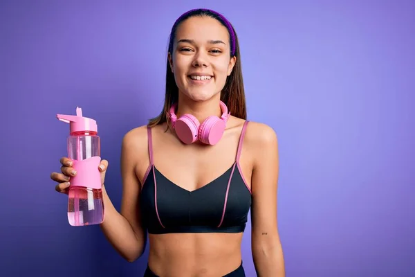 漂亮的女运动员用耳机做运动 喝着一瓶水 面带微笑地站在那里 脸上带着自信的笑容 露出牙齿 — 图库照片