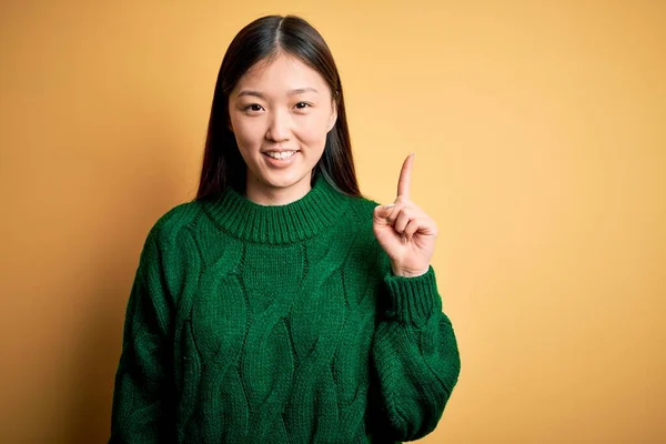 黄色の隔離された背景に緑の冬のセーターを着て若い美しいアジアの女性は 成功したアイデアで指を指しています 興奮し 第一番 — ストック写真