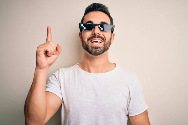 Young handsome man with beard wearing funny thug life sunglasses over white background pointing finger up with successful idea. Exited and happy. Number one.