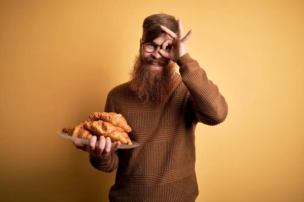 Redhead Irish man with beard eating french croissant pastry over yellow background with happy face smiling doing ok sign with hand on eye looking through fingers