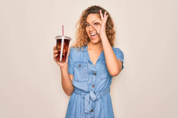 Beautiful blonde woman with blue eyes drinking cola beverage using straw to refreshment with happy face smiling doing ok sign with hand on eye looking through fingers