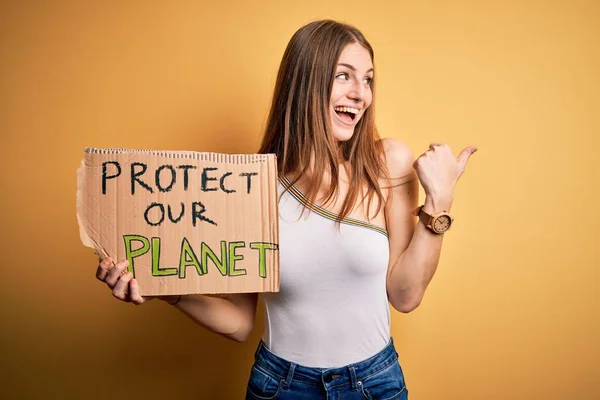 Young redhead woman asking for enviroment holding banner with protect planet message pointing and showing with thumb up to the side with happy face smiling