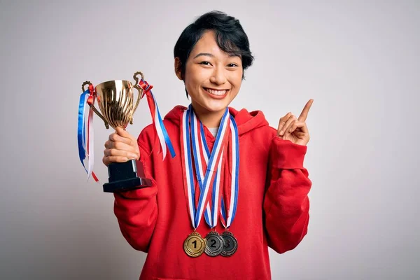 Young beautiful asian girl winner holding trophy wearing medals over white background surprised with an idea or question pointing finger with happy face, number one