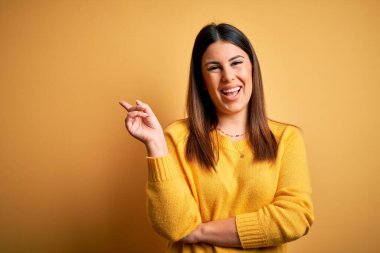 Sarı izole edilmiş arka plan üzerine günlük süveter giyen genç ve güzel bir kadın. Yüzünde kocaman bir gülümseme, el ve parmak ile kameraya bakıyor..