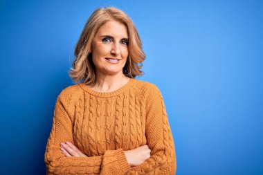 Orta yaşlı güzel sarışın kadın sarı arka planda rahat süveter giyip kollarını kavuşturup kameraya bakarken gülümsüyor. Pozitif kişi.