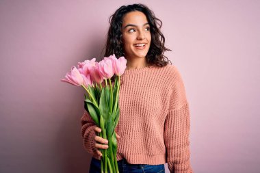 Kıvırcık saçlı, genç ve romantik bir kadın elinde pembe lalelerle yüzünde gülümsemeyle, doğal bir ifadeyle yan yana bakıyor. Kendinden emin gülen.