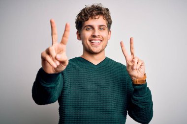 Sarışın, kıvırcık saçlı, yeşil süveter giyen, beyaz arka planda gülümseyen ve parmaklarını zafer işareti yapan genç bir adam. İki numara..