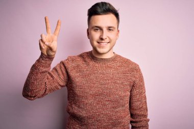 Pembe izole edilmiş arka planda sıradan bir kazak giyen yakışıklı beyaz adam kameraya bakıp zafer işareti yaparken gülümsüyor. İki numara..