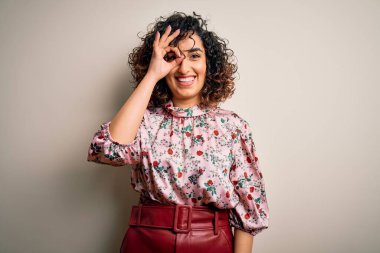 Genç ve güzel kıvırcık Arap kadın çiçekli tişört giyiyor. İzole edilmiş beyaz arka planda duruyor. El hareketleriyle gülümsüyor..