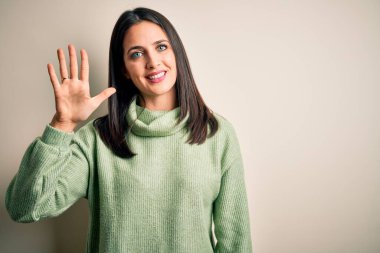 Mavi gözlü, balıkçı yaka kazak giyen genç esmer kadın beyaz arka planda beş numarayı gösterirken kendinden emin ve mutlu bir şekilde gülümsüyor..
