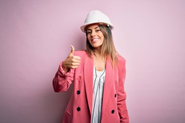 Genç, güzel, esmer mimar kadın pembe arka planda güvenlik kaskı takıyor ve el hareketleriyle mutlu mesut hareket ediyor. Kameraya bakıp başarıyı gösteren ifade onaylanıyor.