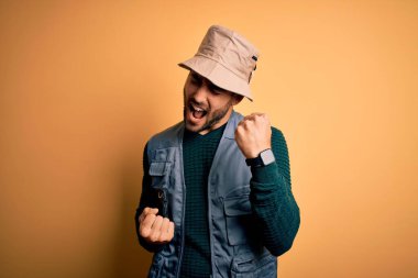 Tatilde sakallı yakışıklı turist sarı arka planda kaşif şapkası takıyor. Kolları havada ve gözleri kapalı olarak başarıyı kutluyor ve şaşırıyor. Kazanan konsept.