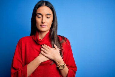 Genç, güzel esmer kadın, izole edilmiş mavi arka planda kırmızı bir süveter giyiyor. Elleri göğsünde, gözleri kapalı ve yüzünde minnet dolu bir jest ile gülümsüyor. Sağlık kavramı.
