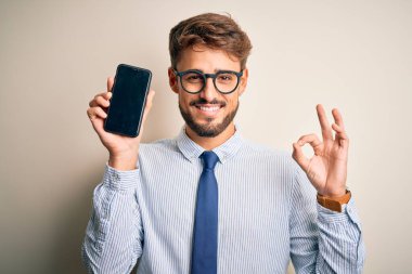 Akıllı telefonu elinde tutan genç işadamı izole edilmiş beyaz arka planda duruyor. Tamam işareti yapıyor. Parmaklarıyla, mükemmel bir sembol.