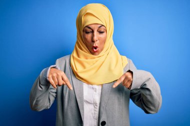 Orta yaşlı esmer iş kadını, geleneksel Müslüman tesettüre bürünmüş mavi arka planda parmaklarını göstererek reklamını yapıyor, şaşırmış yüzünü ve ağzını açıyor.