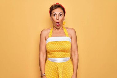 Orta yaşlı bir kadın, 50 'li yaşlarda, sarı arka planda eski tarz bir elbise giyiyor. Şaşırmış ve şaşırmış bir ifade, korku ve heyecanlı bir yüz ifadesi var..