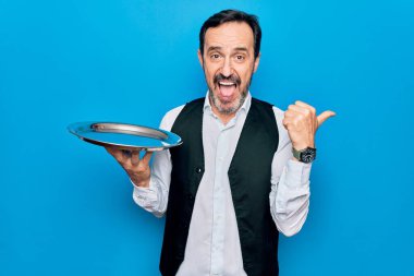 Orta yaşlı, yakışıklı garson adam, mavi arka planda duran tepsiyi tutarken başparmağını yukarı kaldırıyor ve ağzı açık gülümsüyor.