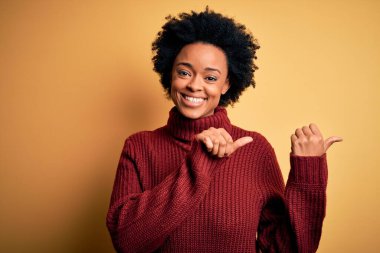 Kıvırcık saçlı, balıkçı yaka kazak giyen, elleri ve başparmakları havada, kendinden emin gülümseyen güzel Afro-Amerikan kadın.