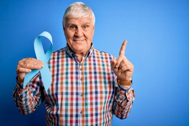 Üst düzey yakışıklı, elinde mavi kanser kurdelesi olan bir adam izole bir arka plan üzerinde bir fikir ya da soru işareti ile şaşırmış, mutlu yüzlü bir numara,