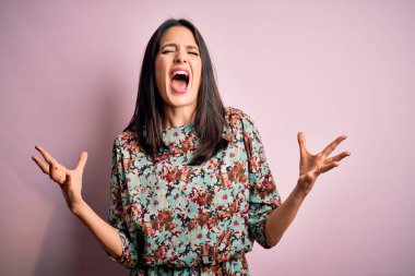 Mavi gözlü genç esmer kadın pembe arka planda çiçekli elbiseler giyiyor deli deli deli bağırıyor ve agresif bir ifade ve kollarla bağırıyor. Hayal kırıklığı konsepti.