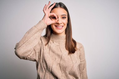 Genç, güzel, esmer, sıradan bir süveter giyen, beyaz arka plan üzerinde duran, el hareketleriyle gülümseyen, mutlu yüzüyle parmaklarının arasından bakan bir kadın..
