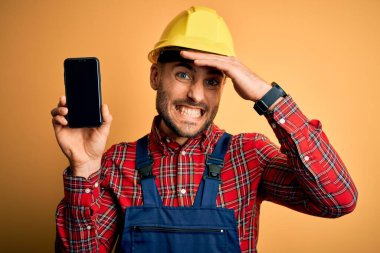 Güvenlik kaskı takan genç müteahhit sarı arka planda akıllı telefon ekranı gösteriyor. Elleri başının üstünde, utanç ve şaşkınlık dolu, sinirli ve hayal kırıklığına uğramış bir halde. Hata için korku ve üzüntü.