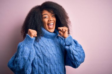 Pembe arka planda kışın süveter giyen Afro saçlı güzel Afro-Amerikalı kadın kollarını kaldırıp, gözleri açık bir şekilde başarısını kutluyor. Kazanan konsept.