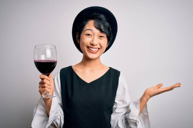 Genç, güzel Asyalı şarap garsonu kız izole edilmiş beyaz arka planda bir bardak kırmızı şarap içiyor mutlu ve heyecanlı, kazanan yüz ifadesi zaferi büyük bir gülümsemeyle kutluyor ve ellerini kaldırıyor.