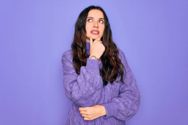 Genç ve güzel bir kadın, balıkçı yaka kazak giyip mor arka planda dikiliyor. Bir soru hakkında endişeleniyor. Endişeli ve gergin.