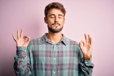 Sakallı, sıradan bir gömlek giyen, pembe arka planda duran, gözleri kapalı gülümseyen ve parmaklarıyla meditasyon hareketi yapan genç yakışıklı bir adam. Yoga konsepti.