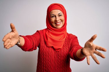 Orta yaşlı esmer kadın izole edilmiş beyaz arka plan üzerine geleneksel tesettür takmış kameraya bakarak sarılmak için kollarını açmış gülümsüyor. Mutluluğu kucaklayan neşeli bir ifade..